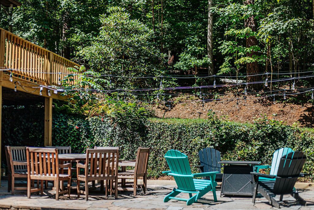 An outdoor patio with wooden furniture and Adirondack chairs, surrounded by dense greenery and trees.