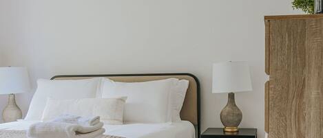 En pænt redt seng med hvide sengetøj og et beige tæppe, et natbord med en lampe og et trænatbord.