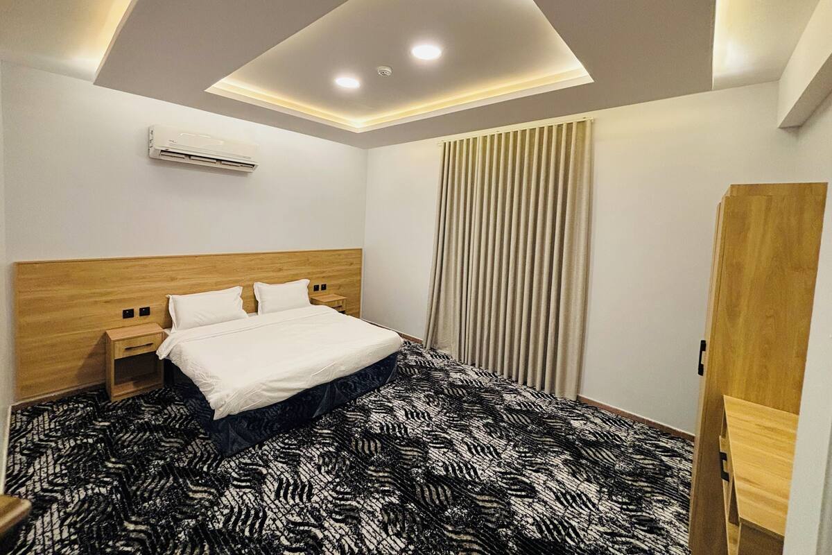 A modern hotel room with a wooden headboard, a bed with white linens, a patterned carpet, and a ceiling light fixture.
