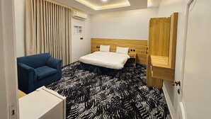 Hotellihuone, jossa on sänky, sininen nojatuoli, kuvioitu matto, kattovalaisin ja puinen pääty.