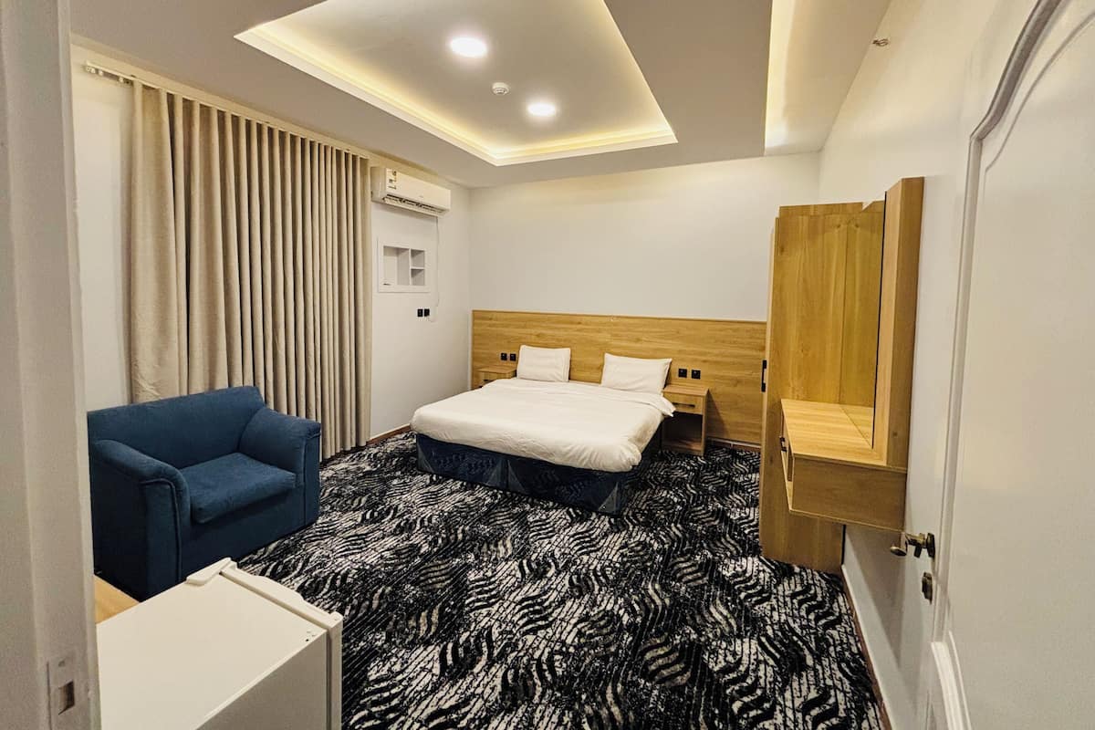 A hotel room with a bed, a blue armchair, a patterned carpet, a ceiling light, and a wooden headboard.
