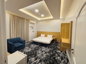 A hotel room with a bed, a blue armchair, a patterned carpet, a ceiling light, and a wooden headboard. - اونارا ستاي 2 للشقق المخدومه (Madinah)
