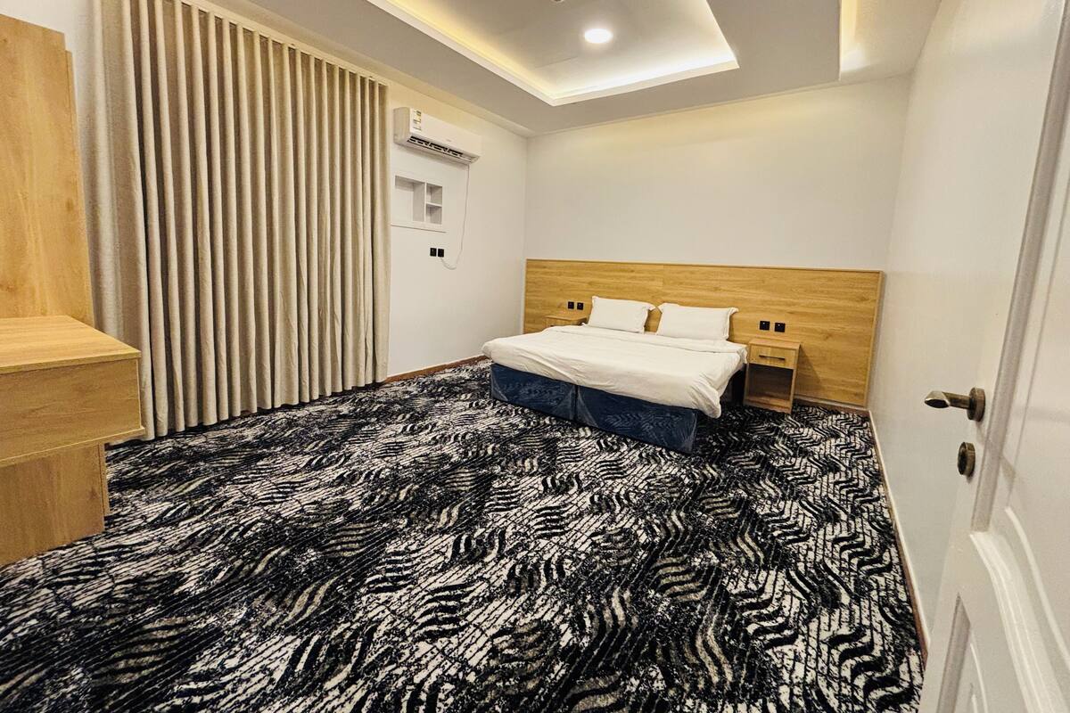 A hotel room with a bed, a desk, a wardrobe, a ceiling light, and an air conditioning unit.