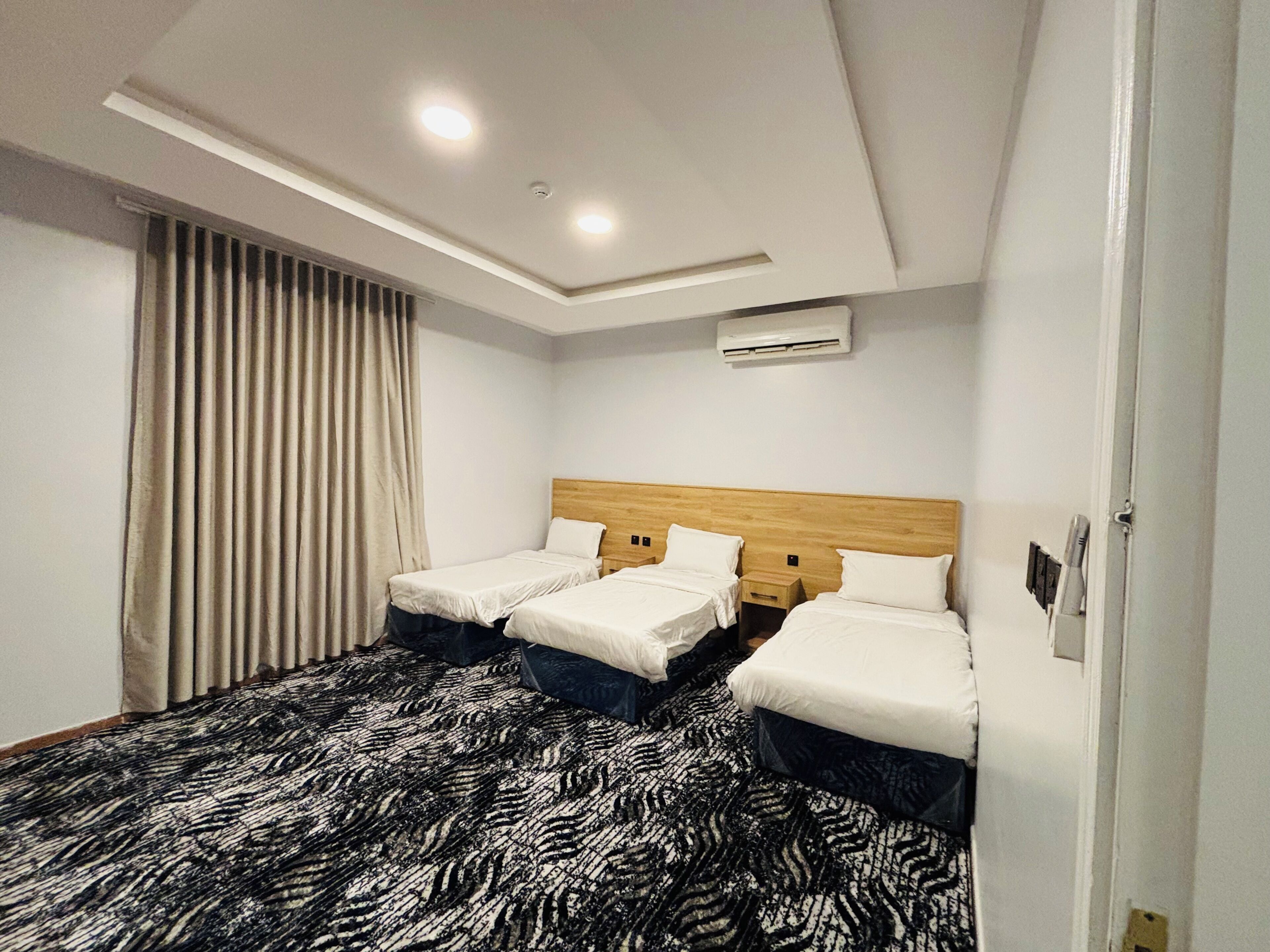 A hotel room with two beds, a patterned carpet, a ceiling light, and an air conditioning unit.