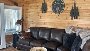Et hyggeligt sommerhusinteriør med en lædersofa, et rundt sofabord og en hund, der hviler på sofaen.