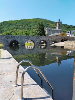 A stone bridge over a calm body of water with a reflection, a metal railing, and a hill with greenery in the background.