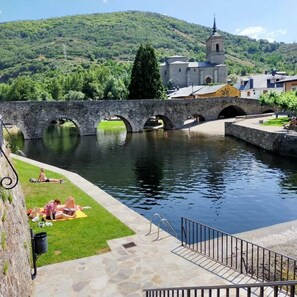 A stone bridge over a calm water body with a grassy area and a building with a tower in the background.