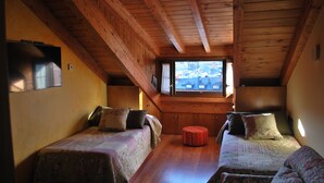 Una habitación con vigas de madera, dos camas con ropa de cama estampada, un pequeño reposapiés y una ventana con vistas a edificios.