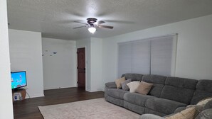 Un soggiorno con un divano grigio, un tappeto beige, una ventola a soffitto e una televisione.