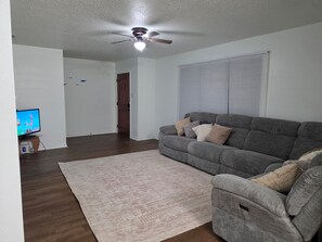 Ein Wohnzimmer mit grauem Sofa, einem beigefarbenen Teppich, einem Deckenventilator und einem Fernseher.