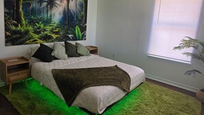 Camera da letto con un grande murale a tema tropicale, un letto con illuminazione verde sottostante e una finestra con persiane.