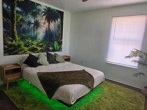 Ein Schlafzimmer mit großem Wandbild im tropischen Stil, einem Bett mit grüner Beleuchtung von unten und einem Fenster mit Jalousien.