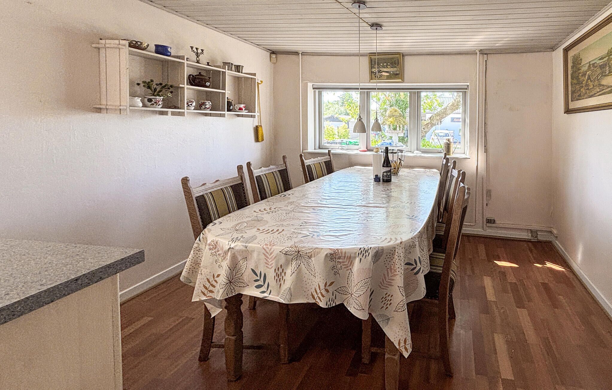 A dining room with a long table, chairs, a window with a view, and shelves with various items.