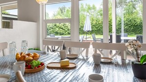 Et spiseområde med et bord dækket til morgenmad, inklusiv croissanter, frugt og ost, med store vinduer der giver udsigt til det fri.