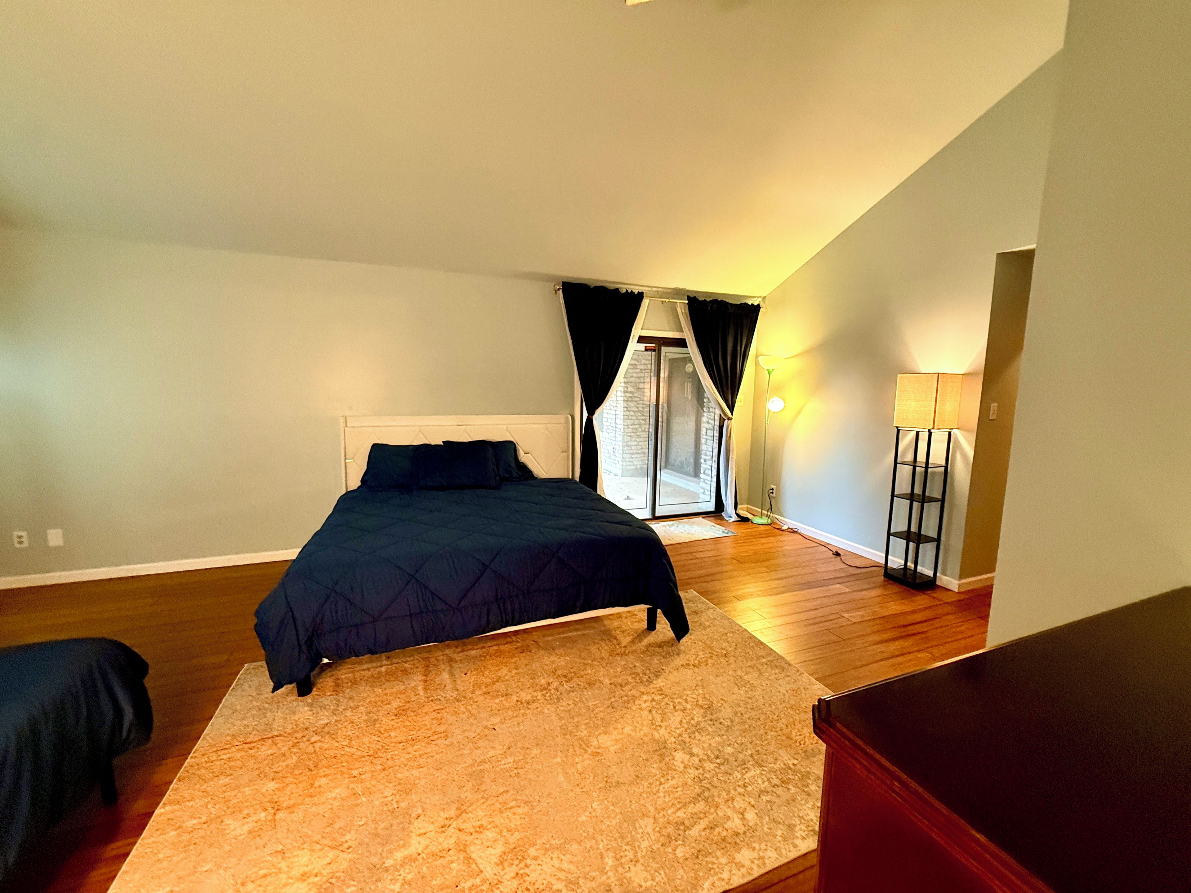 A bedroom with a bed, a nightstand, a lamp, and a view of a balcony.