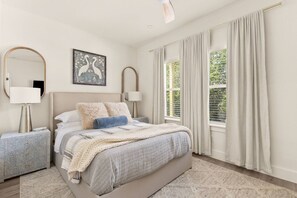 Ein Schlafzimmer mit einem Bett, zwei Nachttischen, einem Wandbild und einem großen Fenster mit Vorhängen.