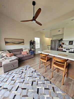 A modern living room with a sofa, a dining area with two chairs, a ceiling fan, and a kitchen with appliances. - Vista Nest | Hill Country Views • Hot Tub Retreat (Canyon Lake)