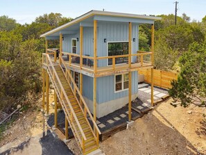 A two-story wooden cabin with a balcony, a staircase, and a fenced yard. - Vista Nest | Hill Country Views • Hot Tub Retreat (Canyon Lake)