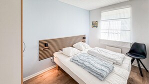 Ein Schlafzimmer mit hölzernem Kopfteil, einem Bett mit weißer und blauer Bettwäsche, einem schwarzen Stuhl und einem Fenster mit durchsichtigen Vorhängen.