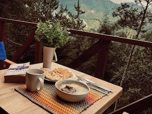 Una mesa con un tazón de sopa, pan plano, una taza, una planta y un libro sobre una terraza de madera con vista a montañas.
