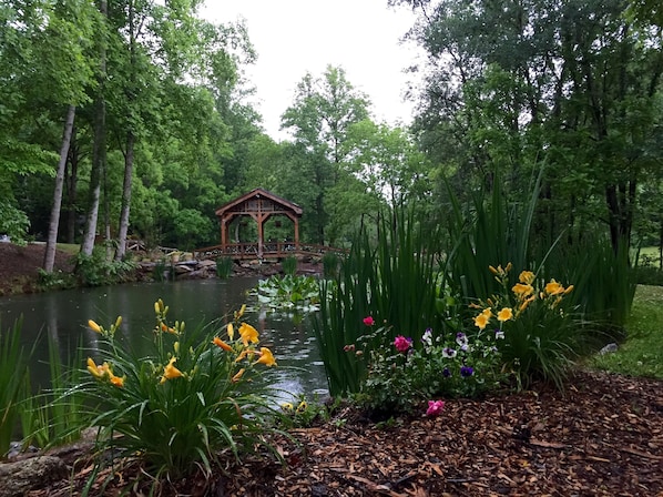 Ein hölzernes Gazebo über einem Teich, umgeben von üppigem Grün und blühenden Pflanzen.