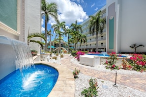 A hotel pool area with a water feature, palm trees, and a pool surrounded by flowers. - Treasure Island Gulf Front Condo Ocean Views (Treasure Island)