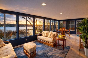 Habitación con un gran ventanal que da a un lago, un sofá, una mesa de comedor con sillas y un taburete pequeño.
