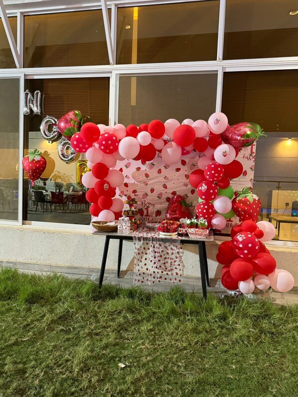 Een tafel versierd met ballonnen en aardbeien, buiten een gebouw met grote ramen.