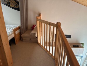 Een slaapkamer met een bed, een klein nachtkastje, een koffer, een houten leuning en een wandplank met een bordje.