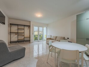 A modern living room with a sofa, a dining table with chairs, a built-in shelving unit, and a sliding glass door leading to a balcony. - Apartment Nala by Interhome (Riva del Garda)