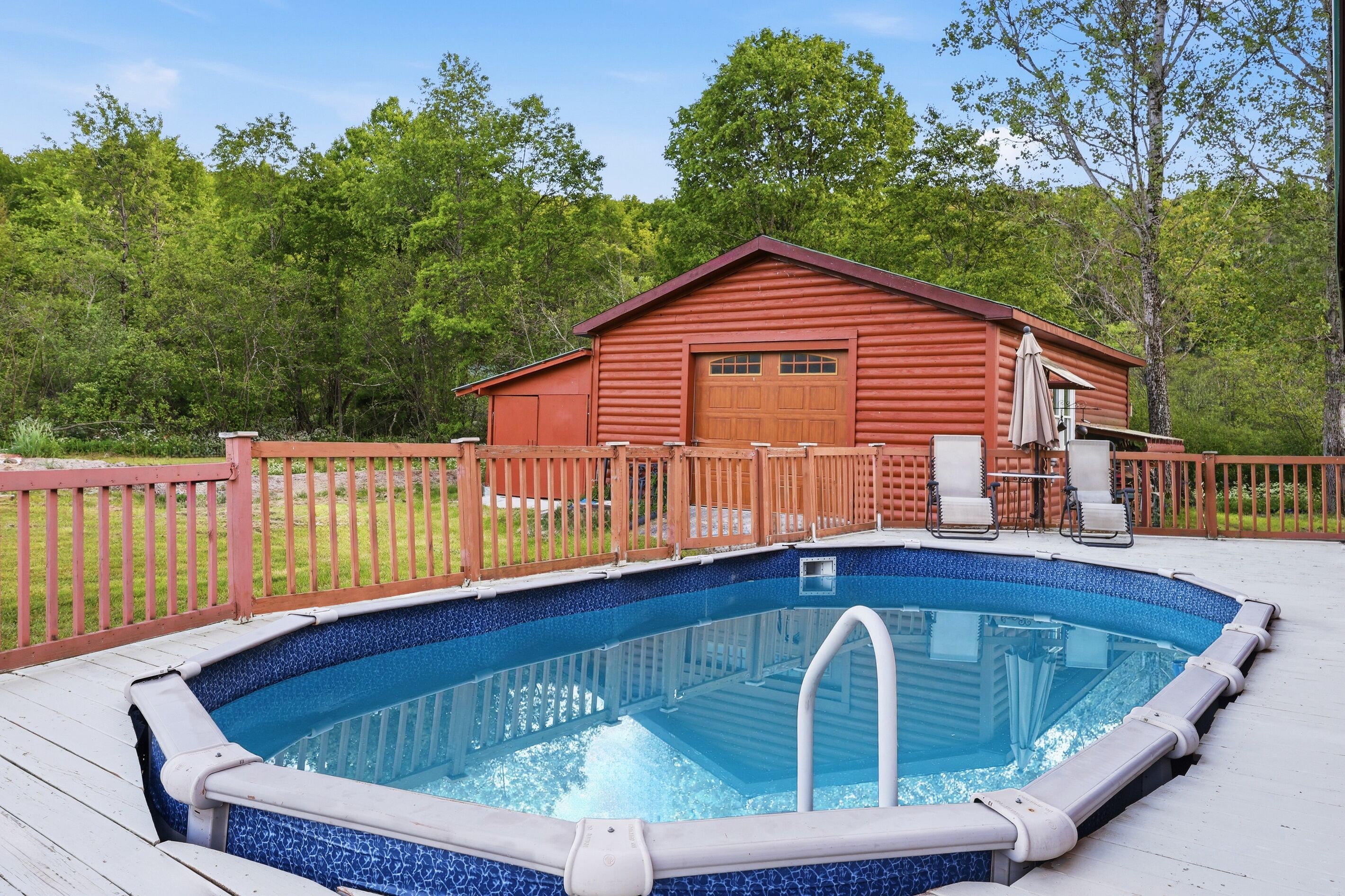 A swimming pool with a wooden deck, a red building with a garage, and a fenced yard.