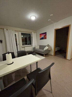 A room with a dining table, chairs, a sofa, a window with curtains, a painting on the wall, and a door leading to another room.