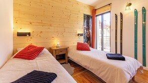 A room with two beds, wooden walls, a window with curtains, a nightstand with a lamp, and skis on the wall. - Le Jovet - AIME LA PLAGNE (AIME LA PLAGNE)