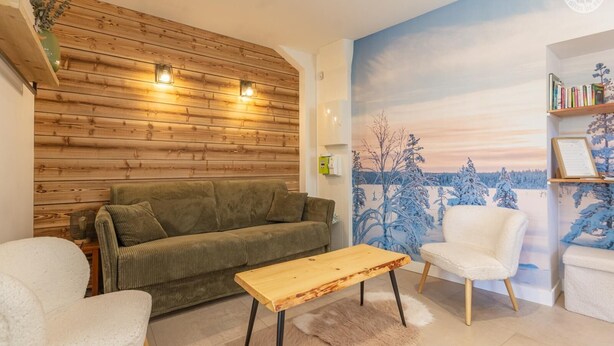 Ein gemütliches Wohnzimmer mit einer Holzwand, einem grünen Sofa, einem weißen Sessel, einem Holztisch und einem Wandgemälde einer verschneiten Landschaft.