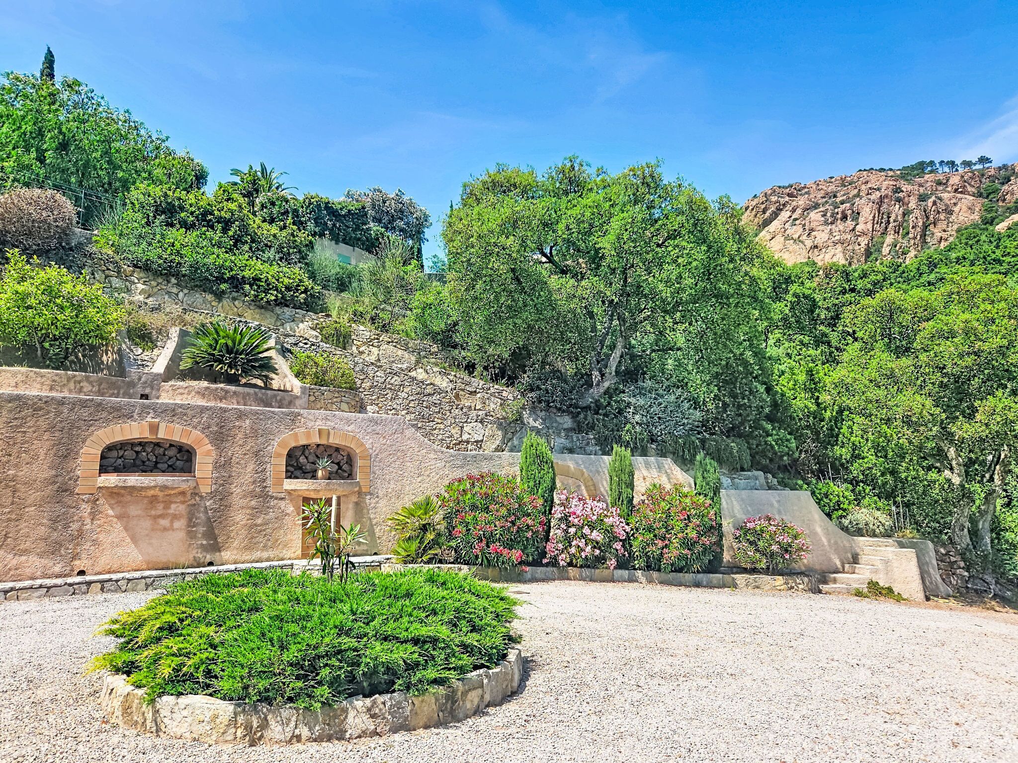 A landscaped area with a gravel path, stone walls, and a variety of greenery including shrubs and flowering plants.