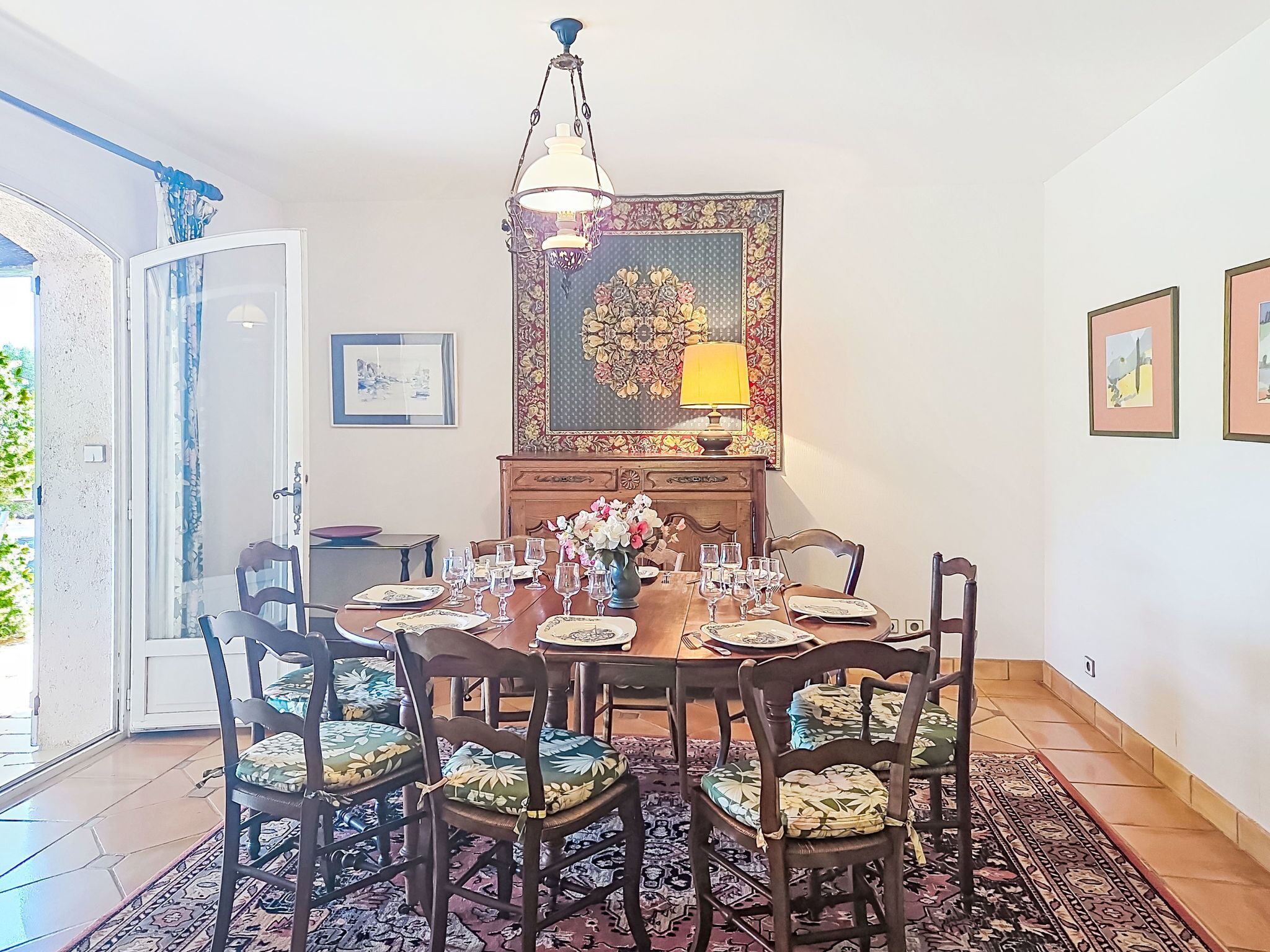 Een eetruimte met een ronde tafel, stoelen, een kroonluchter en een bloemschilderij aan de muur.