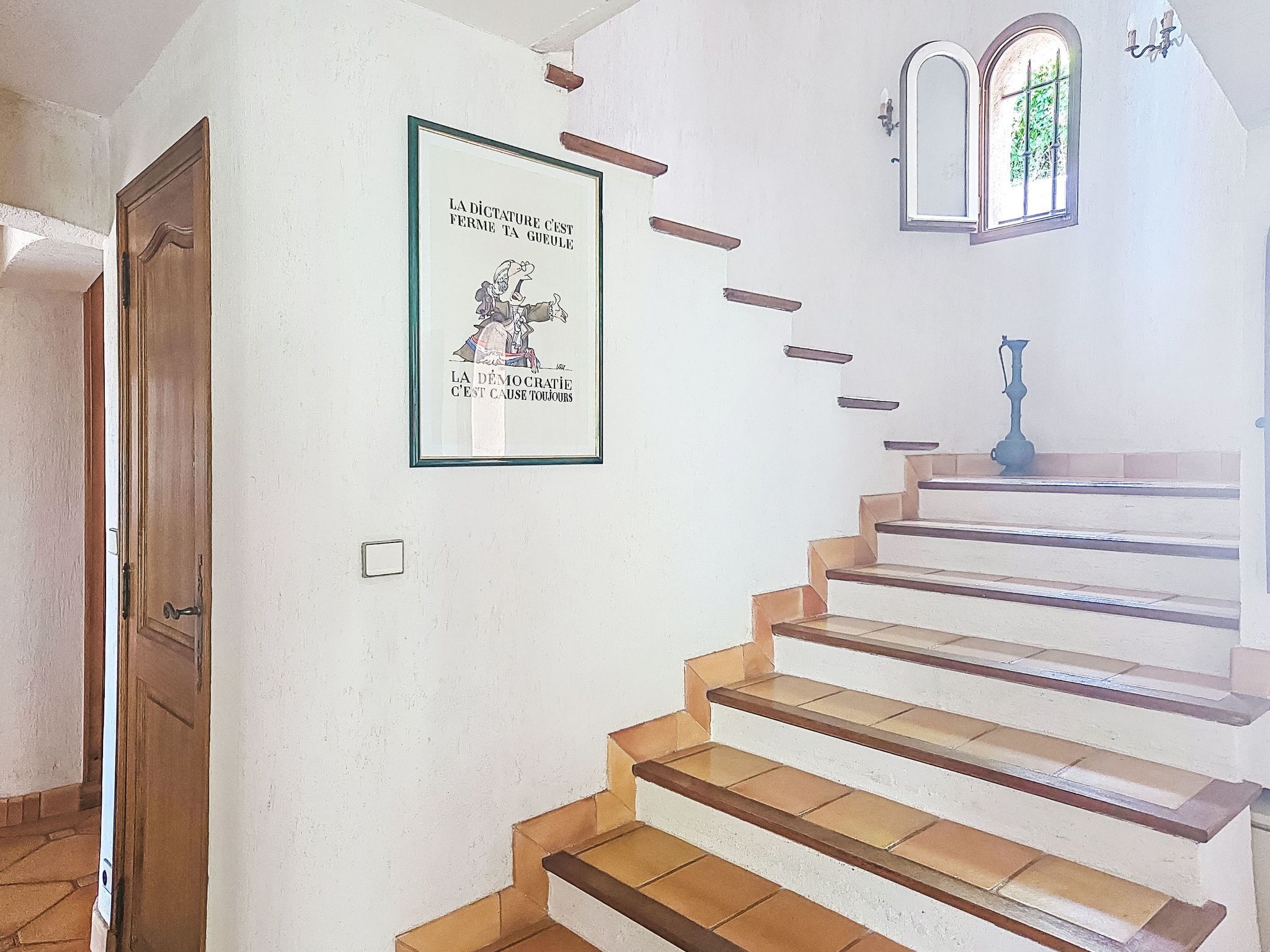 Een trap met een politiek poster in een lijst, een deur en een raam met uitzicht op groen.