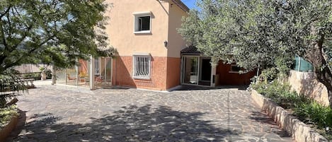 Un bâtiment à deux étages avec une cour pavée de pierres, une petite construction adjacente dotée d’un toit en tuiles et entouré de verdure.