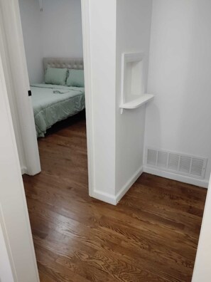 Ein Zimmer mit einem Bett, einer weißen Wand, einem Lüftungsgitter und Holzdielen.