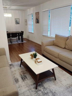 Ein Wohnzimmer mit beiger Couch, einem weißen Couchtisch mit Dekoration, einem Essbereich mit Tisch und Stühlen sowie großen Fenstern mit Jalousien.