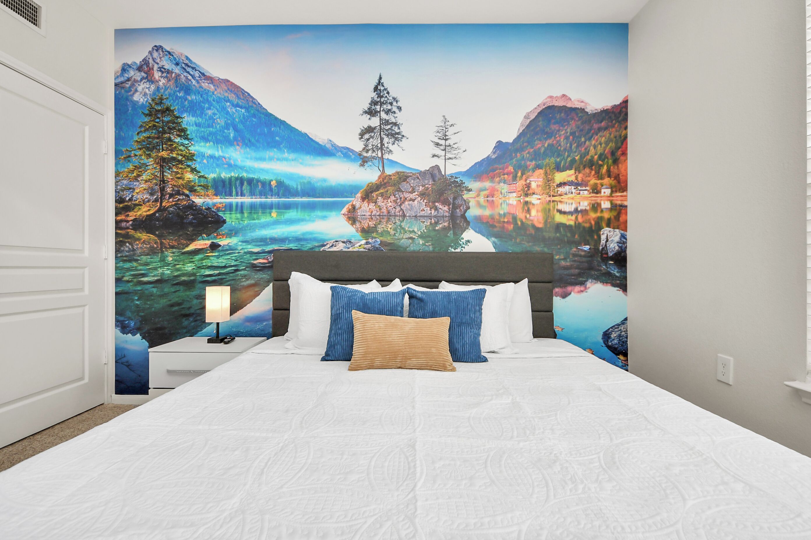 A bedroom with a large bed, a nightstand, a lamp, and a wall mural of a mountain landscape.
