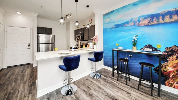 A modern kitchen with a white countertop, stainless steel appliances, and a bar area with stools. - Santorini - 2Bedroom/2Bath/NRG Stadium/Medical Center/Pool/Gym/Sleeps 6 (Houston)