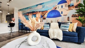 A living room with a blue sofa, a white vase with pampas grass, a painting of a coastal town, and a kitchen in the background. - Santorini - 2Bedroom/2Bath/NRG Stadium/Medical Center/Pool/Gym/Sleeps 6 (Houston)