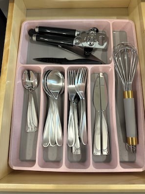 A drawer with a pink interior containing various kitchen utensils including spoons, forks, knives, a whisk, and a grater. - Charming 3-bedroom apartment with AC in vibrant Houston (Houston)