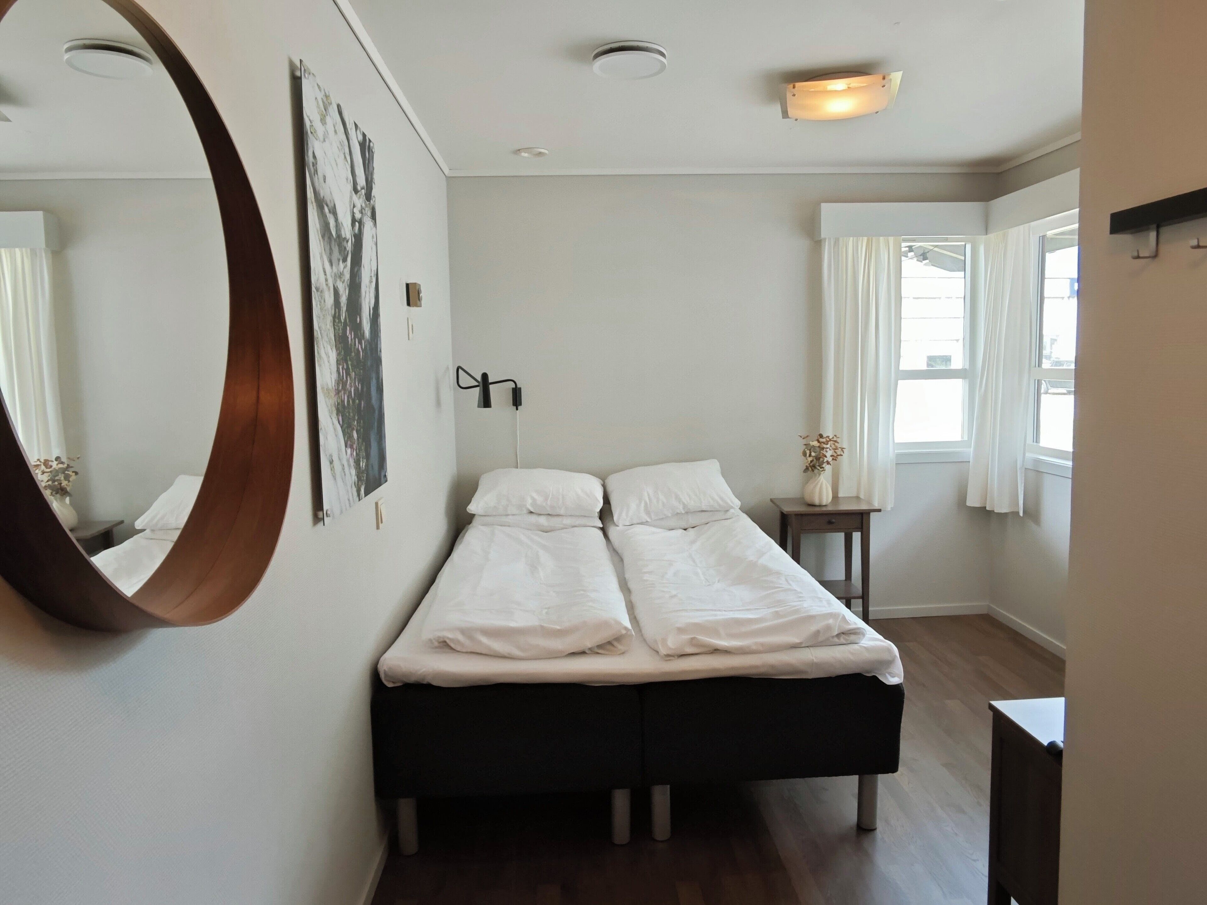 A bedroom with a bed, a bedside table, a mirror, and a window with curtains.