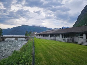 A riverside hotel with a bridge, a grassy lawn, and mountains in the background.