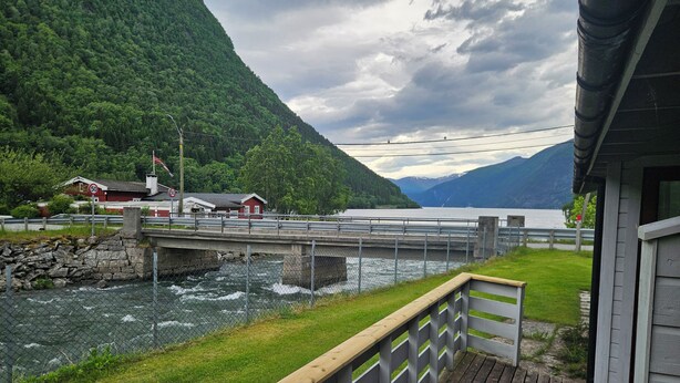 A wooden deck with a railing, a river, a bridge, and a mountain backdrop. - Norddalstunet (Norddal)