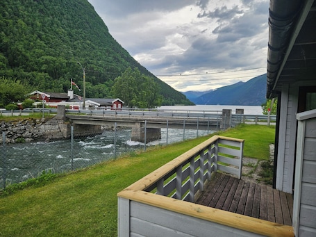 A wooden deck with a railing, a river, a bridge, and a mountain backdrop.. Norddalstunet