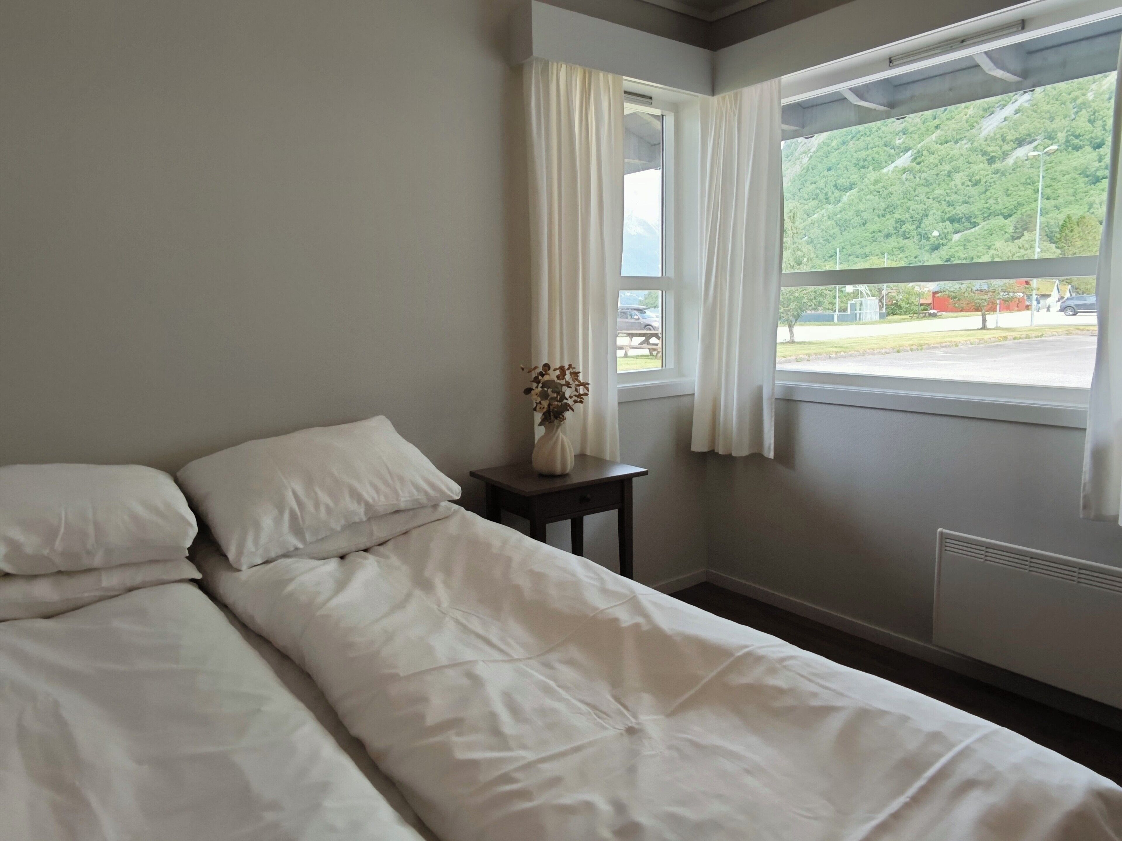 A bedroom with a bed, a nightstand, a vase with flowers, and a window with curtains.