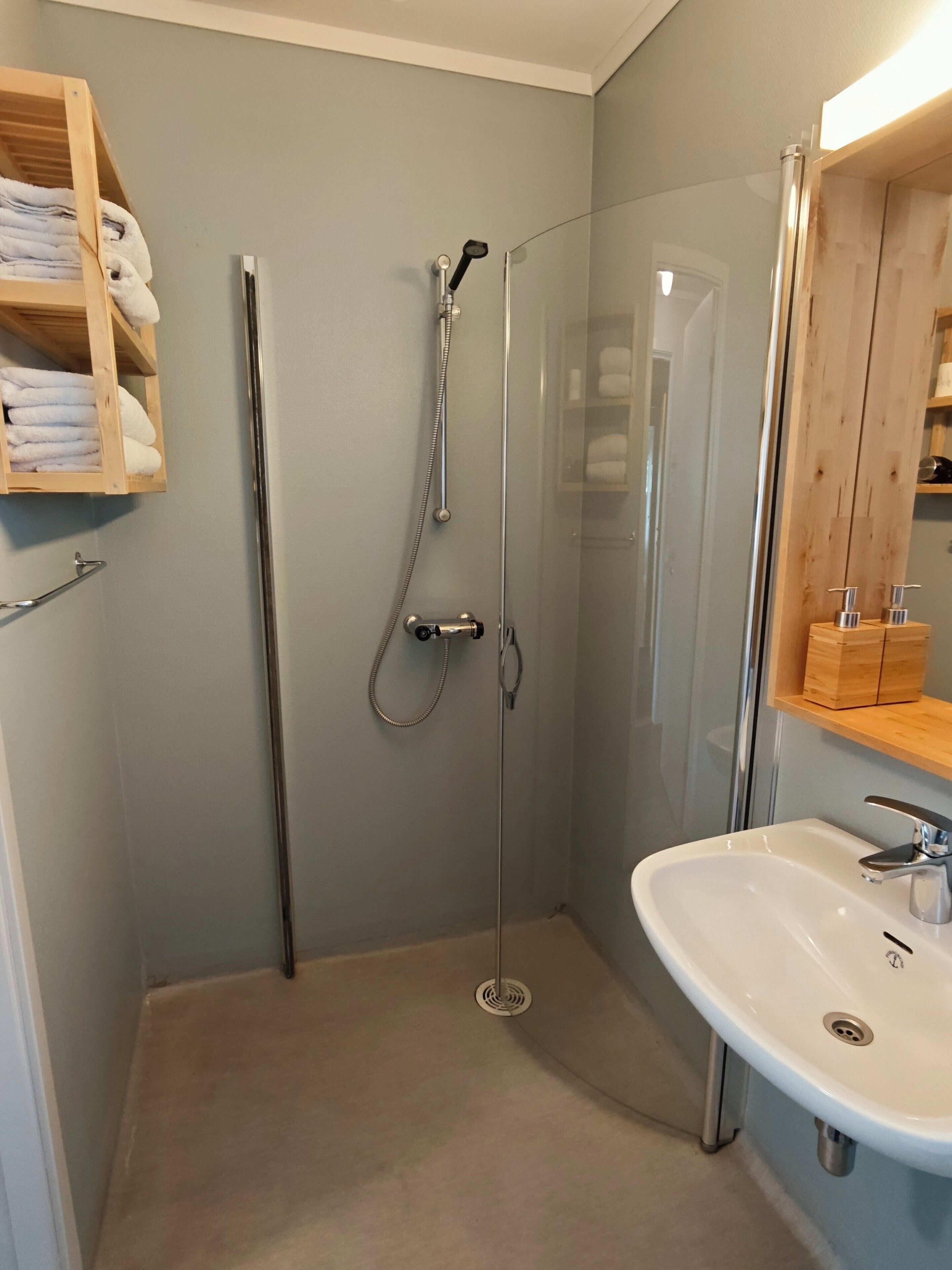 A bathroom with a glass-enclosed shower, a white sink, and a wooden shelf with towels.